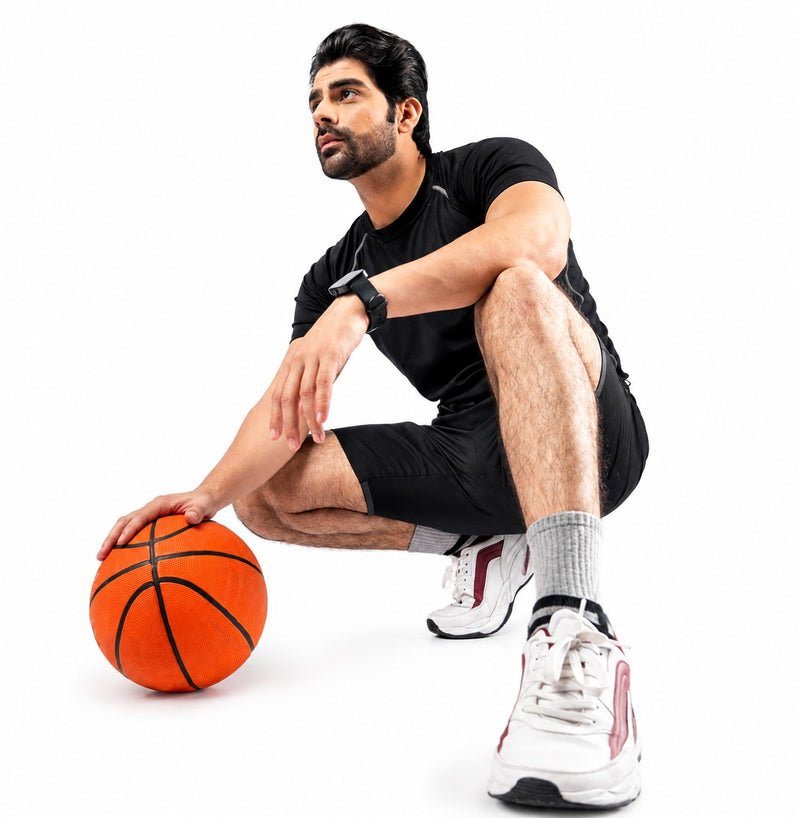 A man in a black athletic t-shirt and shorts crouches on a white studio background while holding an orange basketball. He is wearing white and red sneakers with gray socks.