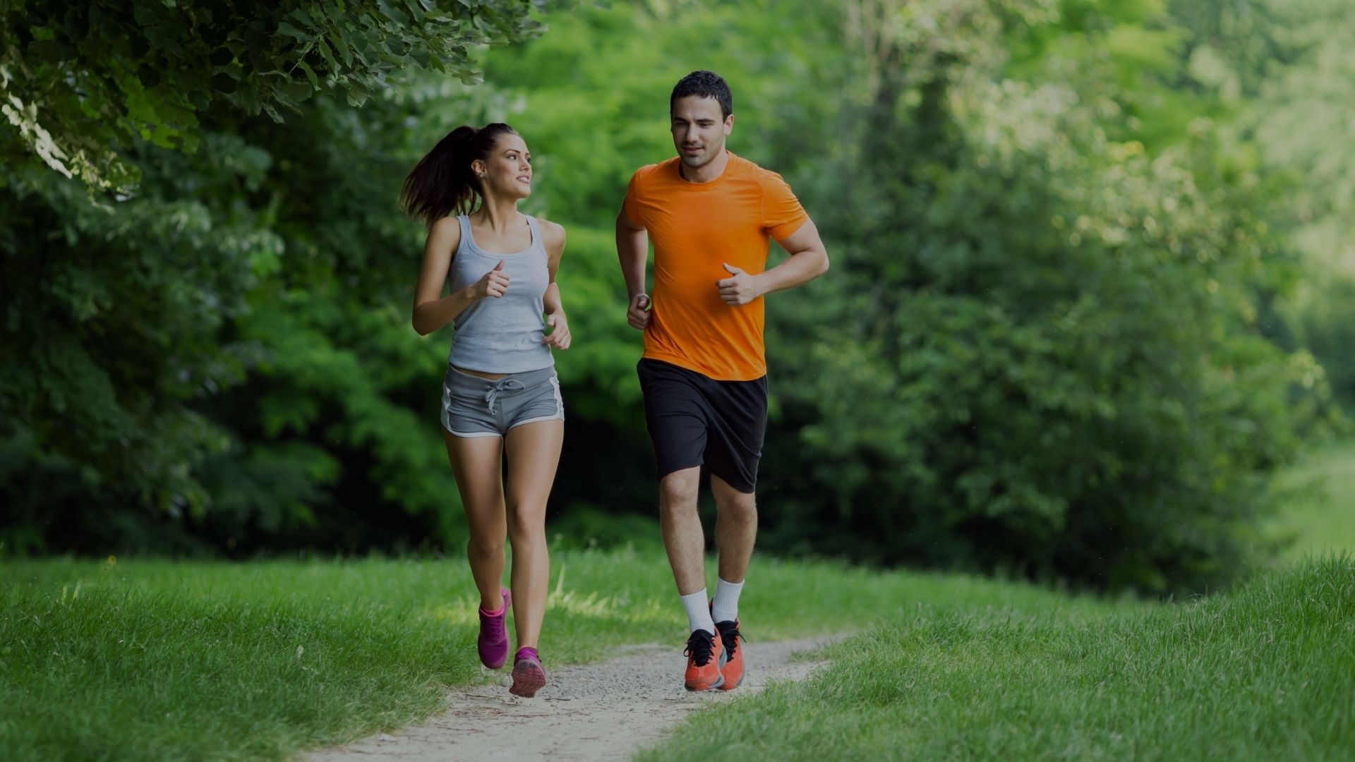 A man and a woman jogging side-by-side on a dirt path in a lush green park. The man is wearing an orange t-shirt, black shorts, and running shoes, while the woman wears a gray tank top, denim shorts, and pink running shoes.
