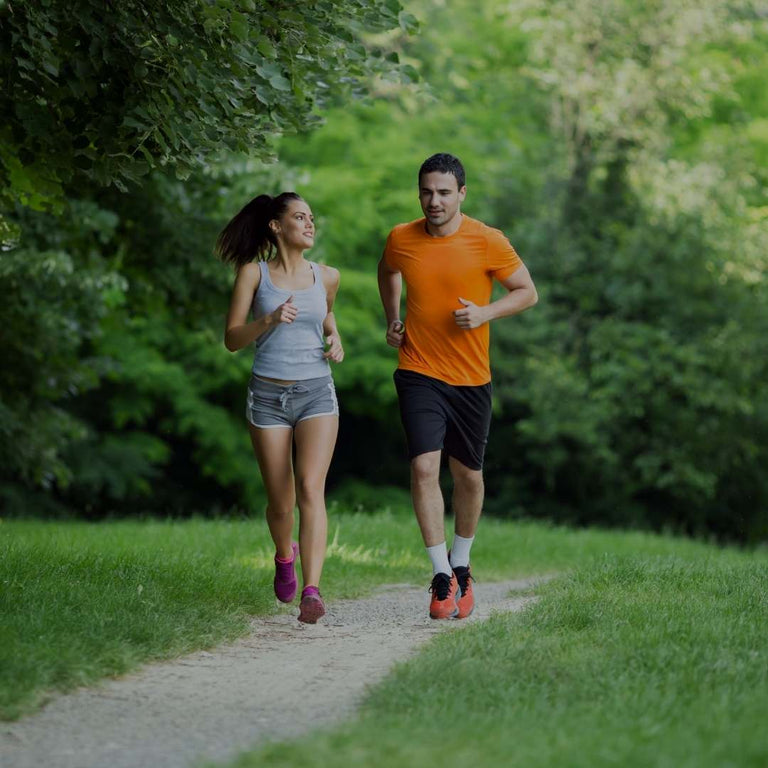 A man and a woman jogging side-by-side on a dirt path in a lush green park. The man is wearing an orange t-shirt, black shorts, and running shoes, while the woman wears a gray tank top, denim shorts, and pink running shoes.