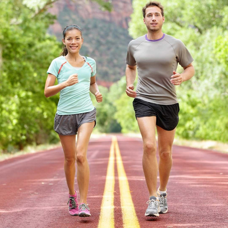 A man and a woman jogging side-by-side on a paved road surrounded by lush green trees, wearing athletic running gear.