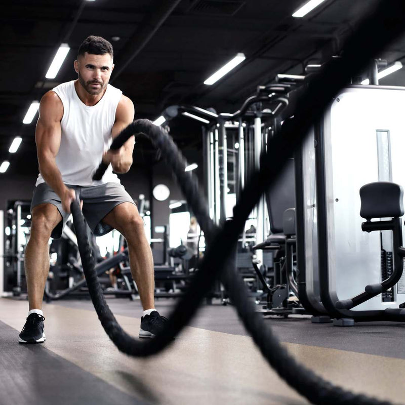 A man in a white tank top and gray shorts performing battle ropes exercises with black battle ropes inside a modern gym.