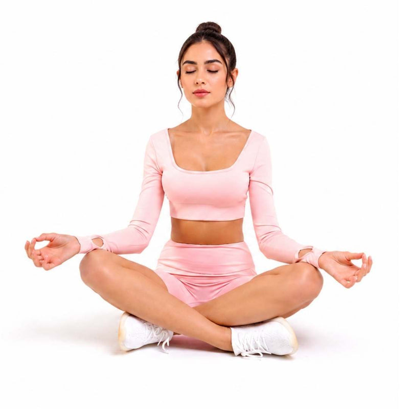 A woman sits cross-legged in a meditative pose wearing a light pink long-sleeve crop top and matching high-waisted shorts, with white sneakers, against a plain white background.